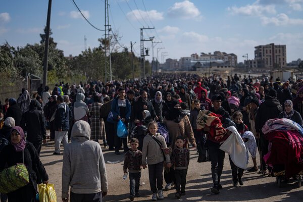 A crowd of people, some of whom carry blankets and other belongings, fills a street. Buildings are seen in the distance.